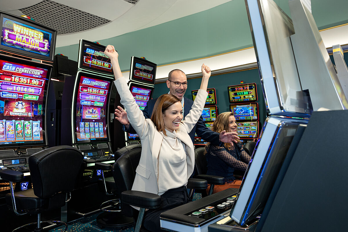 Eine Frau reckt freudig die Arme in die Höhe, während sie vor einem Spielautomaten in einem Casino sitzt. Im Hintergrund sind bunte Bildschirme und andere Gäste zu sehen.