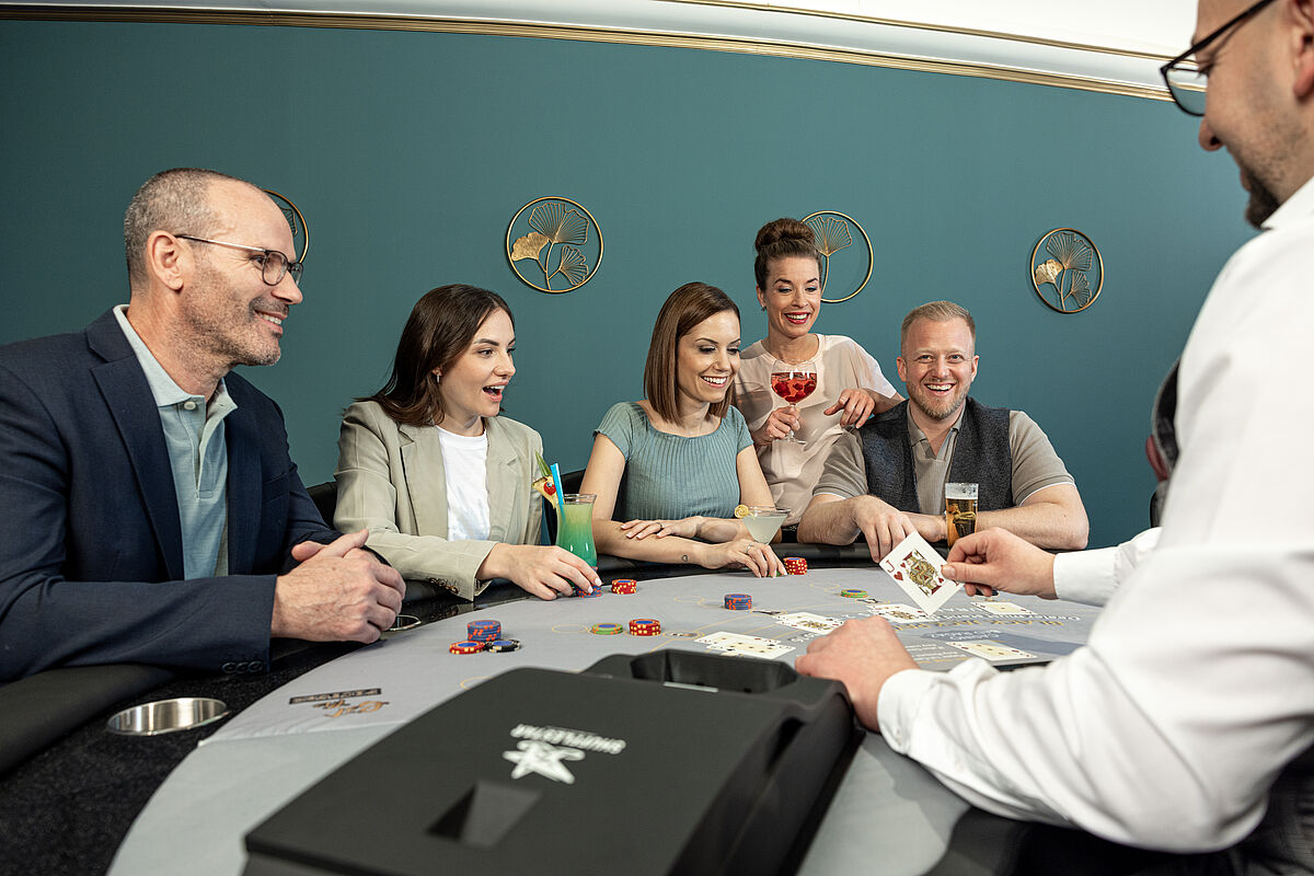 Menschen versammelten sich aufgeregt um einen Pokertisch. Ein Dealer verteilt Karten. Chips liegen verstreut auf dem Tisch und deuten auf ein spannendes Spiel hin.