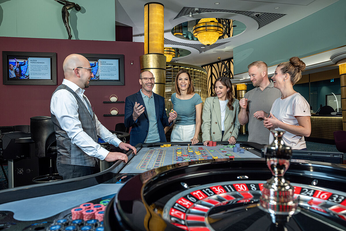 Eine Gruppe von Menschen versammelt sich gespannt um einen Roulettetisch in einem Casino. Ein Croupier leitet das Spiel. Die Stimmung ist lebhaft und gespannt.
